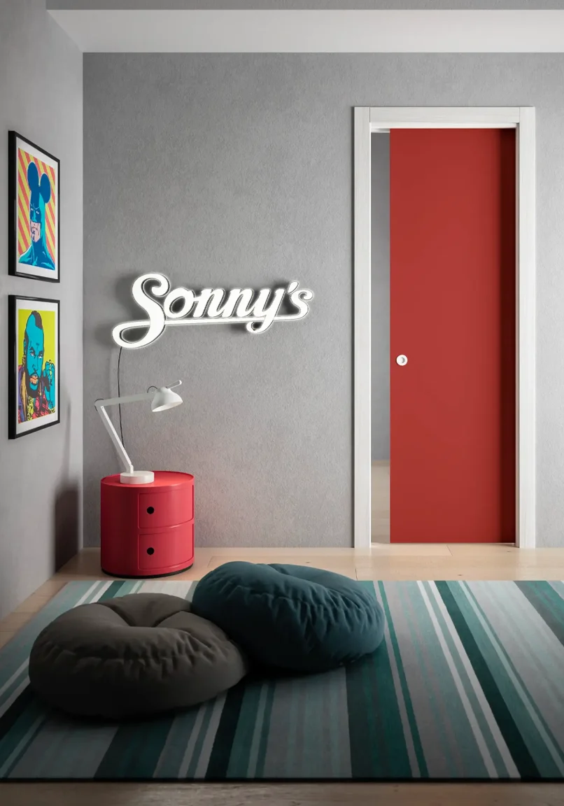 Porta della collezione Cristal scorrevole interno muro con telaio Palissandro bianco e anta in vetro laccato rosso. L'ambiente ha le caratteristiche di una stanza per ragazzi, con super eroi e un tappeto con grandi cuscini per sedersi.
